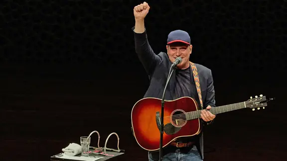 Thees Uhlmann - Der Musiker reckt seinen Arm hoch und steht mit Käppi und Gitarre auf der Bühne der Elbphilharmonie