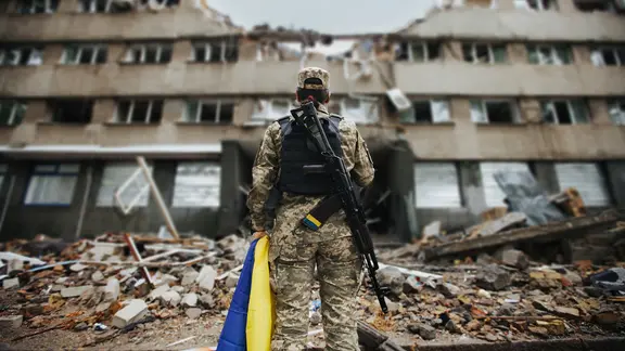 Ukrainische Soldatin mit ukrainischer Flagge in den Händen vor dem Hintergrund eines explodierten Hauses | AdobeStock/alimyakubov Ukrainische Soldatin mit ukrainischer Flagge in den Händen vor dem Hintergrund eines explodierten Hauses