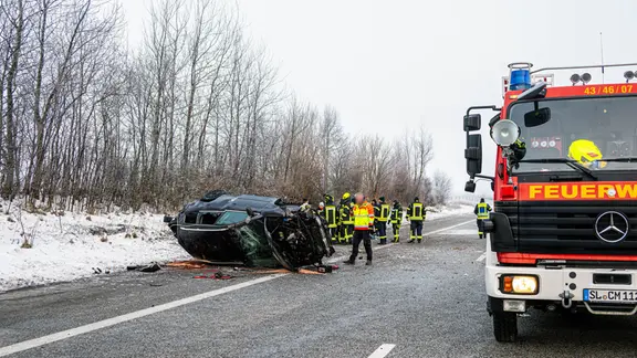 Einsatzkräfte der Feuerwehr stehen auf der Fahrbahn der A7 neben einem zerstörten Pkw nach einem unfall.