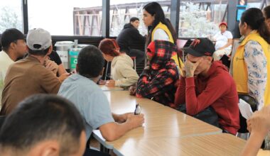 Auf dem Bild sieht man eine Gruppe von Geflüchteten, die an einem hellen Holztisch sitzen. Ein junger Mann mit rotem Pullover vergräbt das Gesicht in den Händen.