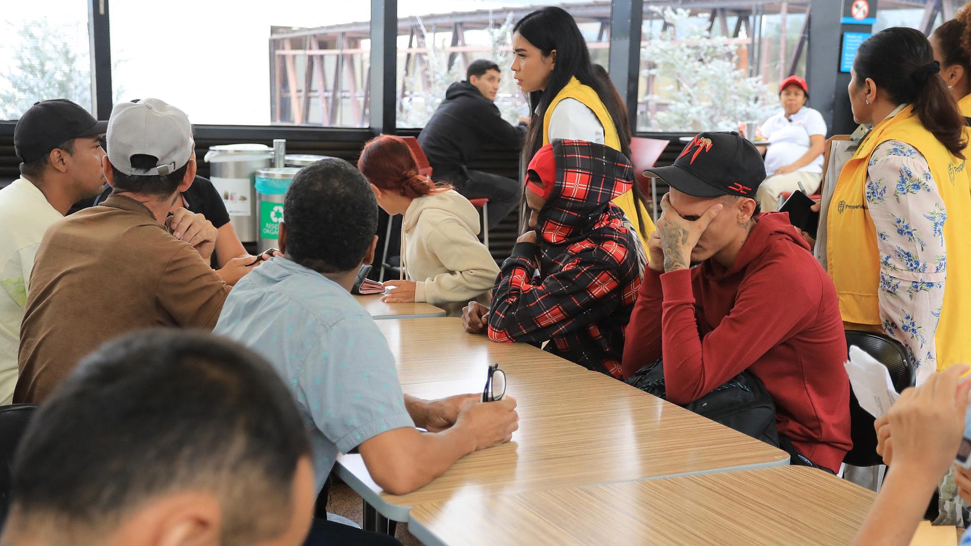 Auf dem Bild sieht man eine Gruppe von Geflüchteten, die an einem hellen Holztisch sitzen. Ein junger Mann mit rotem Pullover vergräbt das Gesicht in den Händen. Auf dem Bild sieht man eine Gruppe von Geflüchteten, die an einem hellen Holztisch sitzen. Ein junger Mann mit rotem Pullover vergräbt das Gesicht in den Händen.