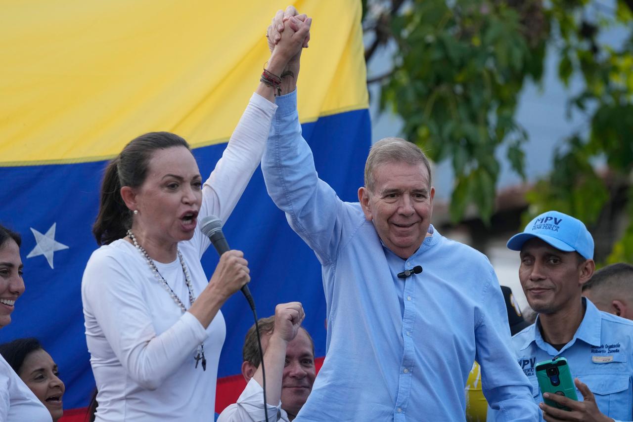Maria Corina Machado und Edmundo Gonzalez Urrutia recken ihre Hände nach oben. Machado hält ein Mikrofon in die Höhe, im Hintergrund die venezolanische Flagge.