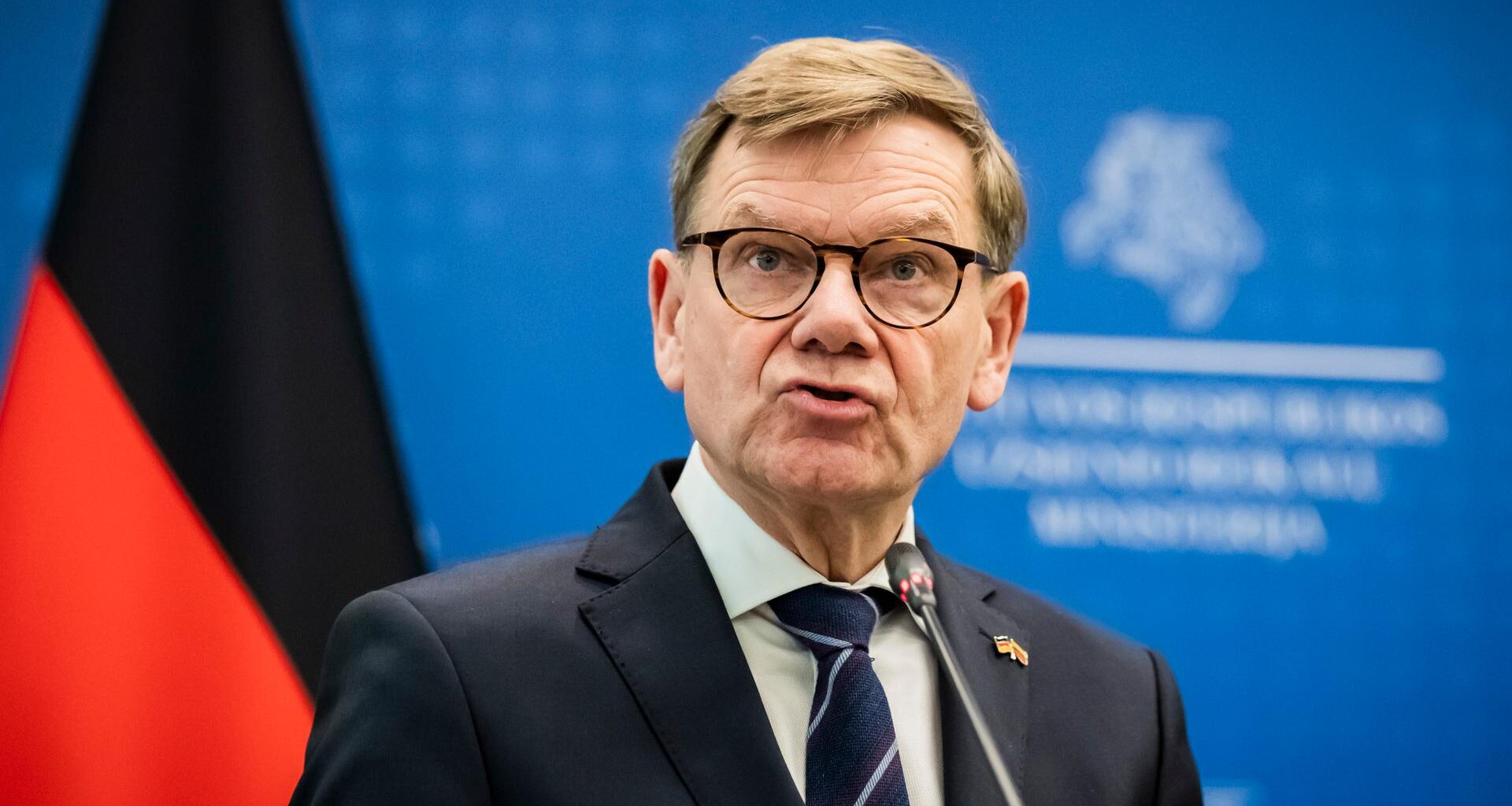 Wadephul steht vor einer blauen Wand mit der deutschen Flagge.