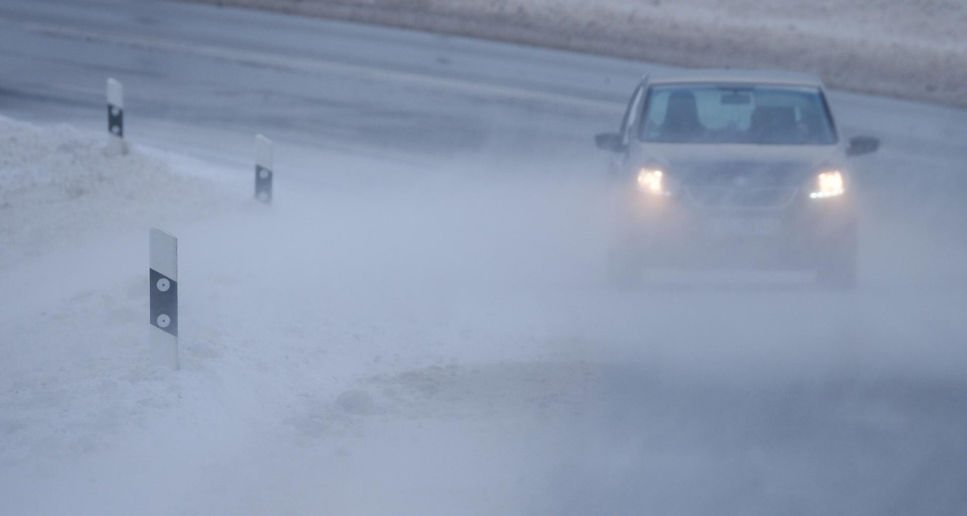 Schnee wird über eine Straße geweht, auf der ein Auto fährt.