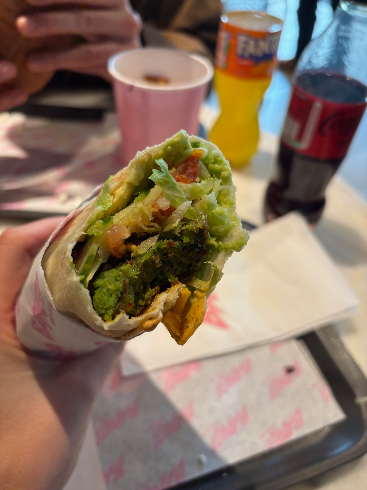 Nahaufnahme eines halb gegessenen Wraps mit Salat, Tomaten, Falafel und Saucen. Im Hintergrund sind Getränke zu sehen.