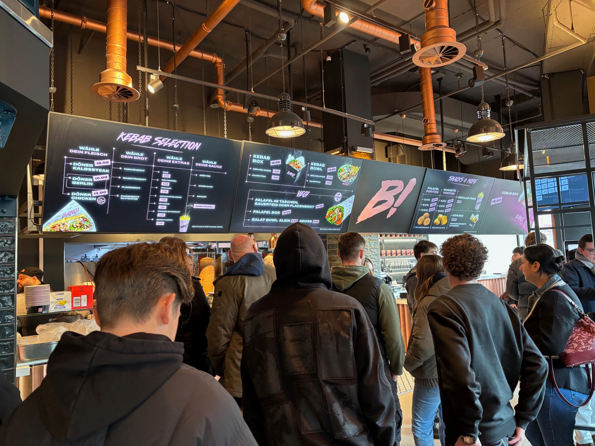 Menschen warten in einer modernen Dönerbude vor einer digitalen Speisekarte mit verschiedenen Kebab-Optionen.