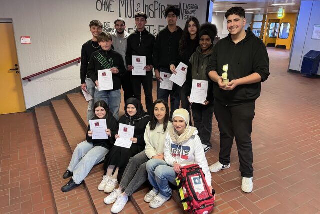 14 Schülerinnen und Schüler an einer Treppe, halten Urkunden, vor einer Schülerin das Erste-Hilfe-Köfferchen. 