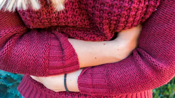 Eine von oben betrachtete Frau im roten Pullover hält sich mit beiden Händen den Bauch.