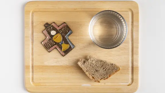 Holzbrett mit einem Kreuz, einem Glas Wasser und einem Stück Brot | picture alliance / imageBROKER | Lucas Seebacher Holzbrett mit einem Kreuz, einem Glas Wasser und einem Stück Brot