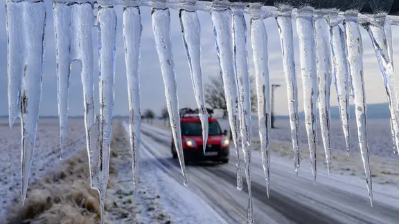 Lange Eiszapfen hängen von einem Verkehrsschild an einer Straße, auf der ein rotes Auto fährt.