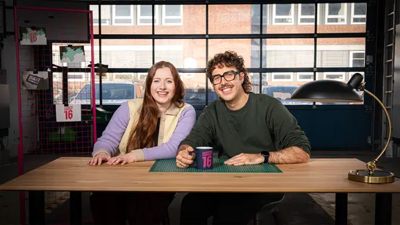 Die Hosts von Studio 16, Anna Albrecht und Jonathan Doll, sitzen an einem Tisch.