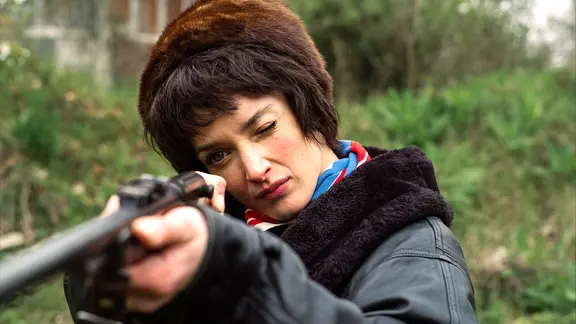 Eine Frau mit Pelzmütze hält eine Flinte abschussbereit hoch, kneift ein Auge beim Zielen zu - Szene aus dem Kinofilm "Niki"