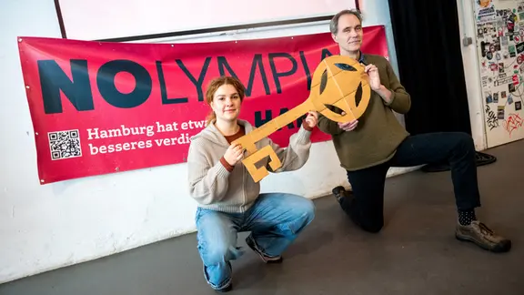 Clara Ihring und Eckart Maudrich von NOlympia Hamburg knieen beim Kampagnenstart gegen die Bewerbung Hamburgs zu Olympischen Spielen vor einem Banner. | picture alliance/dpa | Daniel Bockwoldt Clara Ihring und Eckart Maudrich von NOlympia Hamburg knieen beim Kampagnenstart gegen die Bewerbung Hamburgs zu Olympischen Spielen vor einem Banner.