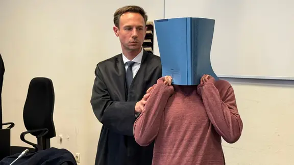 Ein Angeklagter hält sich eine Mappe vors Gesicht, daneben ein Anwalt | NDR/Elke Spanner Ein Angeklagter hält sich eine Mappe vors Gesicht, daneben ein Anwalt