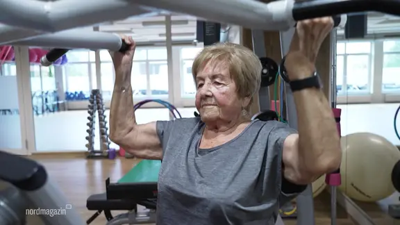 Eine ältere Frau trainiert an einem Fitnessgerät und stemmt zwei Bügel nach oben.