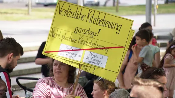 Eine Frau hält ein Schild, auf dem sie verlässliche Kinderbetreuung fordert. | Screenshot Eine Frau hält ein Schild, auf dem sie verlässliche Kinderbetreuung fordert.