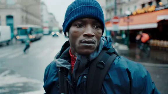 Szene aus dem Film "Souleymanes Geschichte": Ein Mann mit blauer Mütze und dunkler Jacke steht auf einer nassen Straße in der Stadt | Abou Sangaré Szene aus dem Film "Souleymanes Geschichte": Ein Mann mit blauer Mütze und dunkler Jacke steht auf einer nassen Straße in der Stadt