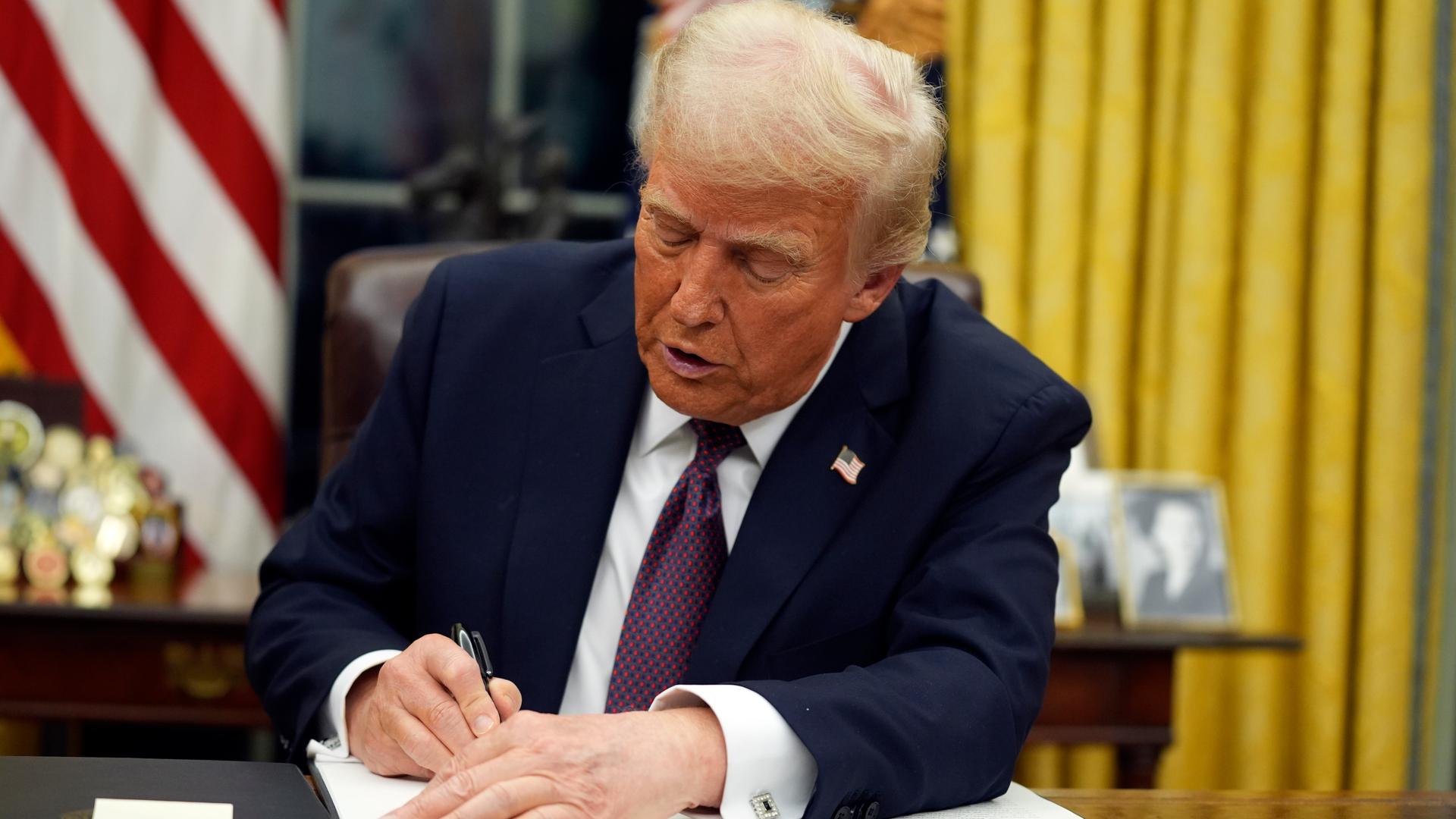 Donald Trump sitzt an einem Schreibtisch vor einer US-Fahne im Weißen Haus und unterzeichnet ein Dekret.
