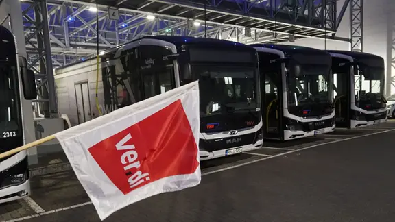 Busse stehen in einem Depot, eine rote ver.di-Fahne wird davorgehalten