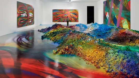 Ausstellungsraum mit bemalten Wänden und einem großen, bunten Hügel aus besprühter Erde auf dem Boden. Der Farbverlauf reicht von Rot über Gelb und Grün bis Blau. Auch der Boden ist farbig besprüht, im Hintergrund hängen abstrakte Gemälde.