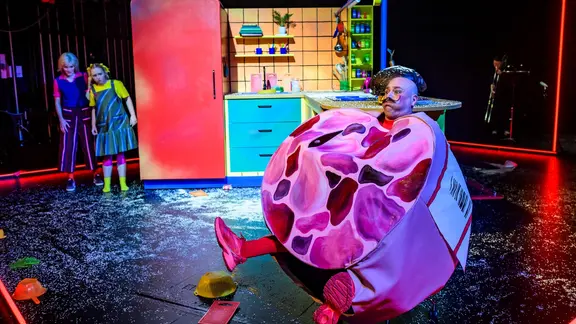 Ein Mann in einem Wurst-Kostüm auf einer Bühne mit zwei Schauspielerinnen | Bettina Stöß Ein Mann in einem Wurst-Kostüm auf einer Bühne mit zwei Schauspielerinnen