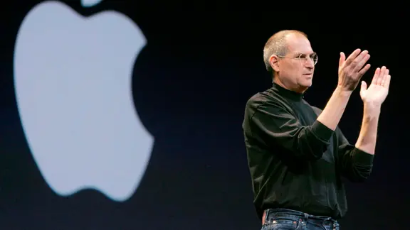 Die Apple-Story - Eine Vision verführt die Welt Die Visionen von Apple-Gründer Steve Jobs haben unsere tägliche Kommunikation und damit unser Lebensgefühl völlig verändert – vor allem durch das iPhone. | © SWR/AP Photo/Paul Sakum Steve Jobs steht auf der Bühne und klatscht. Hinter ihm prangt ein riesiges Apple Logo.