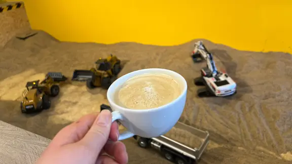 In einem vorgebuchten Zeitfenster kann man an den Geräten spielen und sich Getränke bestellen. | NDR, Larissa Mass Eine Tasse mit Cappuccino vor Baggern im Sand.
