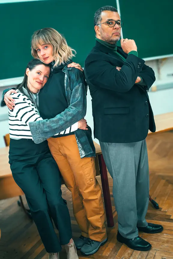 Jennifer Böhm, Tiffany Köberich und Nikolaus Okonkwo in einem Thriller-Kammerspiel. | Struck, Schauspielhaus Kiel In einem Klassenzimmer umarmen sich zwei Frauen. Ein Mann steht mit dem Rücken zu ihnen.