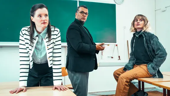 Jennifer Böhm, Nikolaus Okonkwo, Tiffany Köberich | Schauspielhaus Kiel/Struck eine Frau Stützt die Arme auf einen Tisch und schaut ins Publikum, eine junge Frau sitzt auf einem Tisch und zwischen Ihnen feht ein Mann.