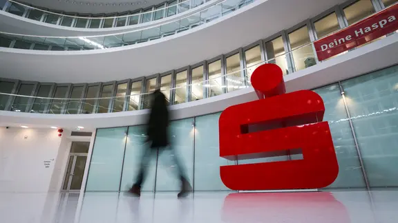 Ein Passant geht an einem großen Sparkassen-Logo vor einer Haspa-Filiale im Deutschlandhaus in der Innenstadt vorbei. | picture alliance/dpa | Christian Charisius, Christian Charisius Ein Passant geht an einem großen Sparkassen-Logo vor einer Haspa-Filiale im Deutschlandhaus in der Innenstadt vorbei.