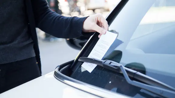 Eine Frau zieht einen Strafzettel hinter dem Scheibenwischer ihres Autos hervor. | dpa, Christin Klose Eine Frau zieht einen Strafzettel hinter dem Scheibenwischer ihres Autos hervor.