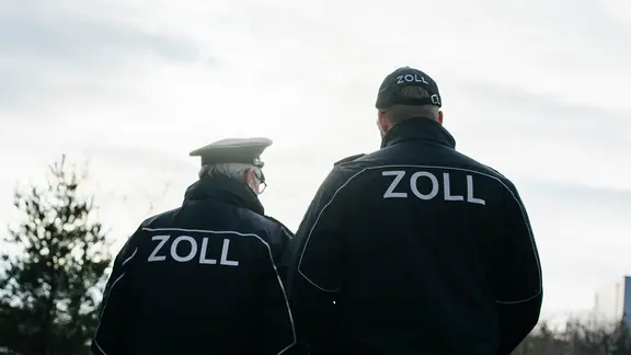 Zwei Männer tragen eine blaue Uniform mit der Aufschrift Zoll auf dem Rücken. | NDR, Julius Matuschik Zwei Männer tragen eine blaue Uniform mit der Aufschrift Zoll auf dem Rücken.