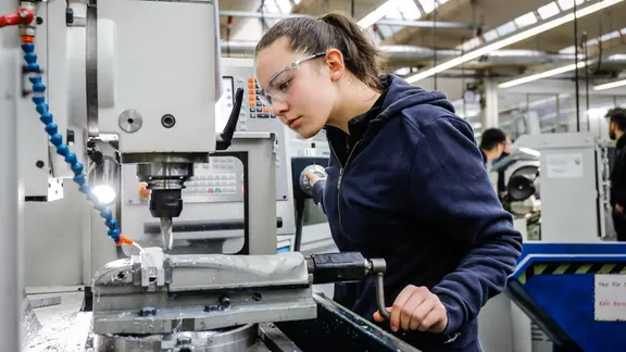 Eine Auszubildende steht mit Schutzbrille an einer Werkzeugmaschine. | picture alliance / Rupert Oberhäuser , Rupert Oberhäuser Eine Auszubildende steht mit Schutzbrille an einer Werkzeugmaschine.