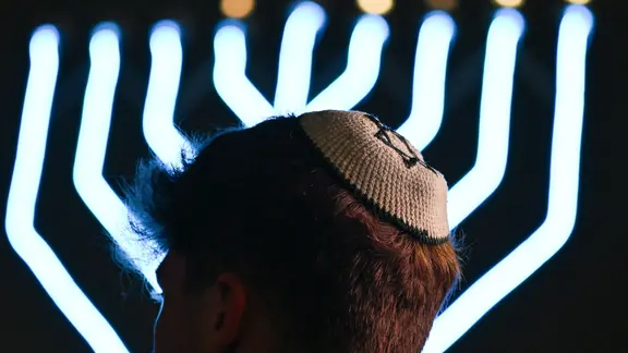 Ein Mann trägt eine Kippa, im Hintergrund leuchtet ein Chanukka-Leuchter. | dpa - picture alliance, Arne Dedert Ein Mann trägt eine Kippa, im Hintergrund leuchtet ein Chanukka-Leuchter.