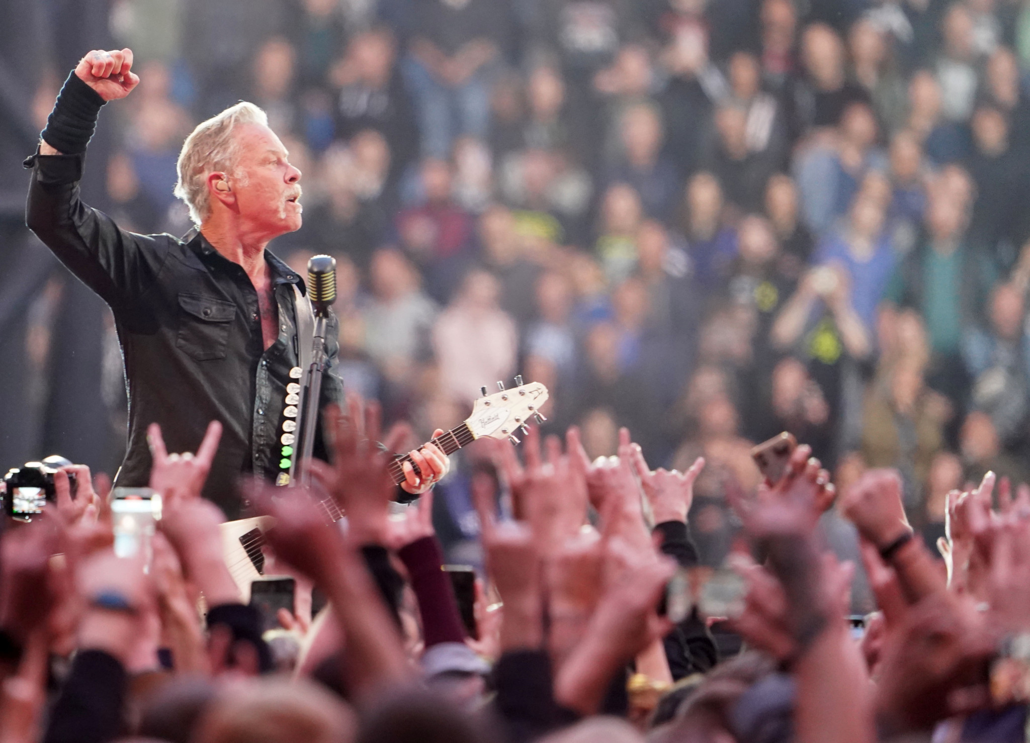 Rockmusiker mit erhobener Faust und Gitarre auf der Bühne vor jubelnder Menschenmenge mit ausgestreckten Händen