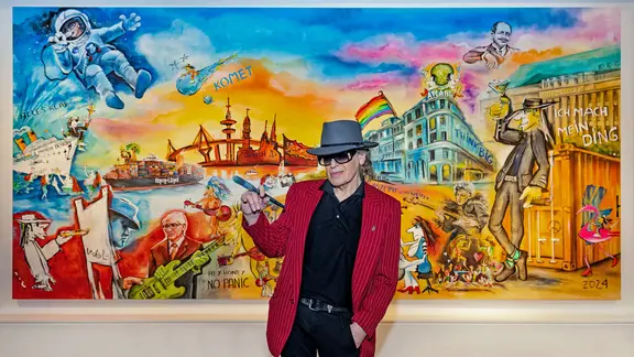 Udo Lindenberg steht mit einem Pinsel in der Hand vor seinem bislang größten Gemälde, das verschiedene Motive aus seiner Musik und der Schifffahrt zeigt. | Tine Acke/Hapag-Lloyd Presseteam /dpa Udo Lindenberg steht mit einem Pinsel in der Hand vor seinem bislang größten Gemälde, das verschiedene Motive aus seiner Musik und der Schifffahrt zeigt.