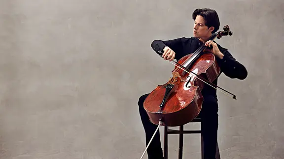 Cellist Daniel Müller-Schott sitzt vor einer grauen Wand und spielt