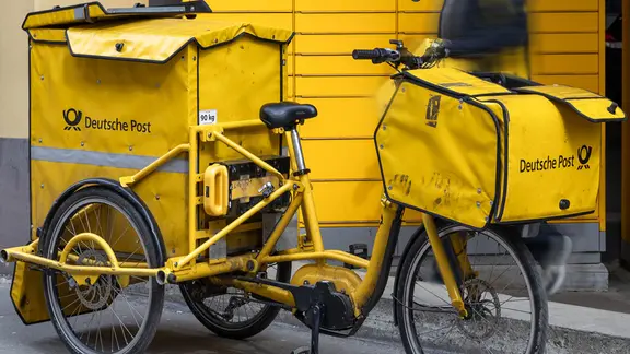 Ein Zustellfahrrad der Deutschen Post steht bereit, während ein Postbote Sendungen zustellt und transportiert. | imago Ein Zustellfahrrad der Deutschen Post steht bereit, während ein Postbote Sendungen zustellt und transportiert.