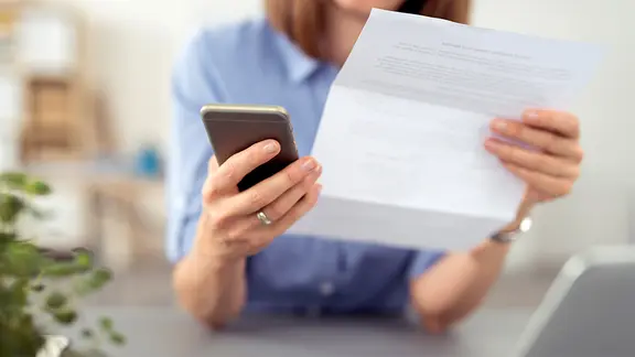 Frau schaut auf einen Brief und tippt auf ihr Mobiltelefon. | Fotolia.com, contrastwerkstatt Frau schaut auf einen Brief und tippt auf ihr Mobiltelefon.