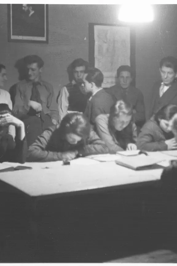 Seit 1927 trafen sich jüdische Jugendliche an verschiedenen Orten und bereiteten sich auf ihre Auswanderung nach Palästina vor. | Stadtgeschichtliches Museum Leipzig/Abram Mittelmann Zionistische Jugendbewegungen, Foto: Atelier Mittelmann. Das Bild zeigt Jugendliche, die sich um einen Tisch herumgruppieren, einzige sitzend, die anderen dahinter stehend. Sie unterhalten sich angeregt oder beugen sich über einige auf dem Tisch ausgebreitete Karten.