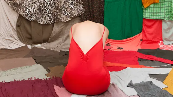 In der Ausstellung "Botenstoffe" dreht sich alles um Textilien. | Sabrina Asche Das Werk "Alle Kleider einer Frau" zeigt einen Frauenkörper von Hinten in einem roten Trägerkleid, aber ohne Kopf, umgeben von zahlreichen bunten Kleidungsstücken.