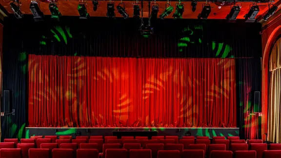 Der Theaterinnenraum der Comödie Lübeck. | Comödie Lübeck GmbH, Comödie Lübeck GmbH Eine Bühne mit einem roten Vorhang, davor stehen viele rote Stühle in verschiedenen Reihen.