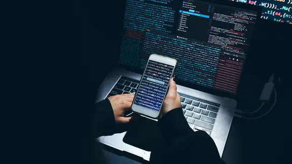 Hände halten ein Smartphone vor einem Laptop. Auf den Bildschirmen befinden sich abstrakte Codes. | photocase.de, przemekklos Hände halten ein Smartphone vor einem Laptop. Auf den Bildschirmen befinden sich abstrakte Codes.
