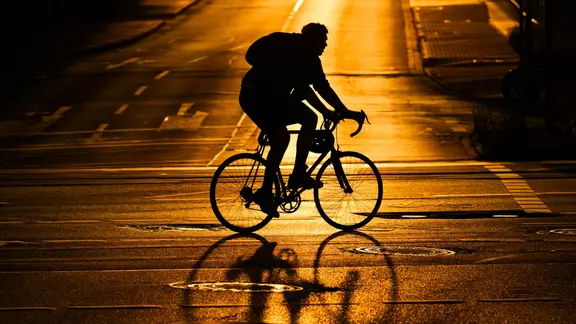 Ein Radfahrer fährt über eine von der Sonne orange angestrahlte Straße | picture alliance/dpa | Julian Stratenschulte Ein Radfahrer fährt über eine von der Sonne orange angestrahlte Straße