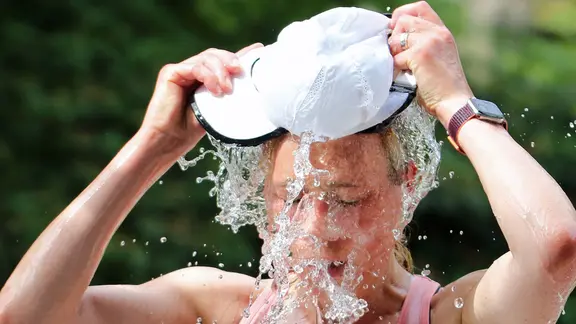 Eine Frau kippt sich Wasser aus einer Mütze über den Kopf.