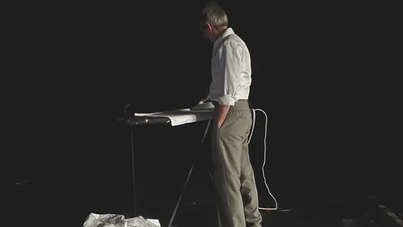 Ein Mann steht auf einer nahezu komplett dunklen Bühne an einem Bügelbrett | Genoveva Allan Ein Mann steht auf einer nahezu komplett dunklen Bühne an einem Bügelbrett