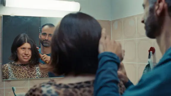 Eine junge Frau steht vor einem Spiegel, ein Mann kämmt ihr die Haare und steht hinter ihr. | realfiction Filmverleih Eine junge Frau steht vor einem Spiegel, ein Mann kämmt ihr die Haare und steht hinter ihr.