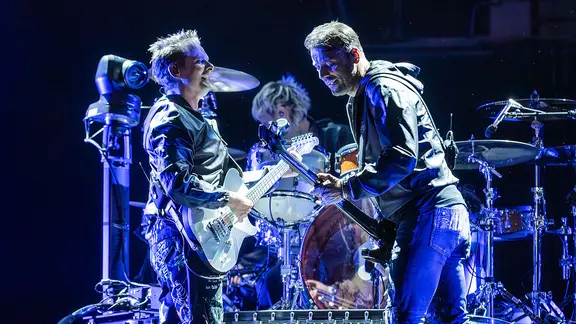 Bandmitglieder von Muse stehen auf der Bühne vor dem Schlagzeug. | picture alliance / IPA | Mirko Fava/TSCK/LiveMedia / ipa- Bandmitglieder von Muse stehen auf der Bühne vor dem Schlagzeug.