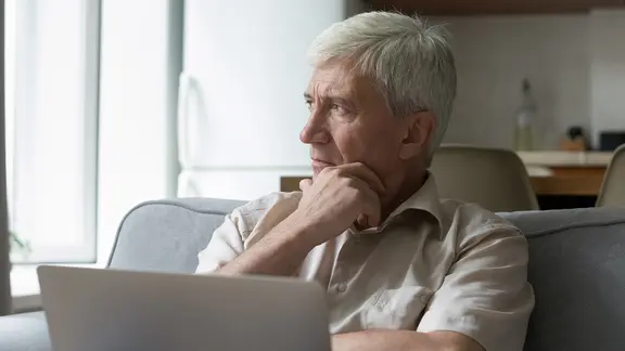 Ein nachdenklicher Mann sitzt zu Hause auf dem Sofa, berührt sein Kinn, blickt vom Computerbildschirm weg und grübelt über einem Geschäftsdokument, um ein Problem online zu lösen. | AdobeStock/fizkes Ein nachdenklicher älterer Mann vor einem Computer