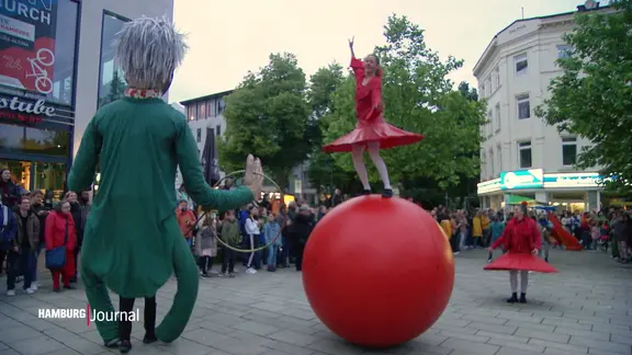 Auf einem öffentlichen Platz schauen viele Menschen einer Tänzerin auf einer großen Kugel und einem Puppenspieler mit einer übergroßen Puppe zu. | Screenshot Auf einem öffentlichen Platz schauen viele Menschen einer Tänzerin auf einer großen Kugel und einem Puppenspieler mit einer übergroßen Puppe zu.